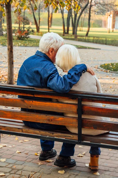 Happy elderly man and woman sitting on a bench in autumn day. Relaxed senior couple sitting on a park bench. Grandfather gently kisses grandmother on the forehead. — Stock Photo, Image