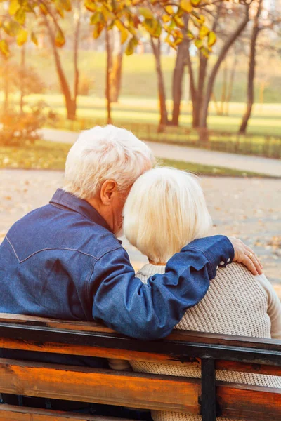 Happy elderly man and woman sitting on a bench in autumn day. Relaxed senior couple sitting on a park bench. Grandfather gently kisses grandmother on the forehead.
