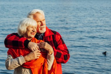 cheerful senior citizens woman and man are standing and hugging on the lake, against the background of the bridge.