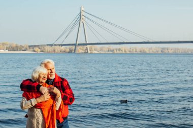 cheerful senior citizens woman and man are standing and hugging on the lake, against the background of the bridge.