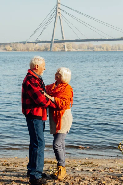 cheerful senior citizens woman and man are standing and hugging on the lake, against the background of the bridge.