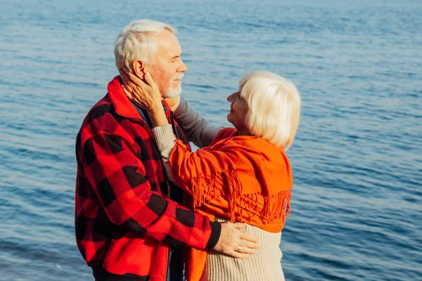 Cheerful senior citizens woman and man are standing and hugging on the lake, against the background of the bridge. — Stock Fotó