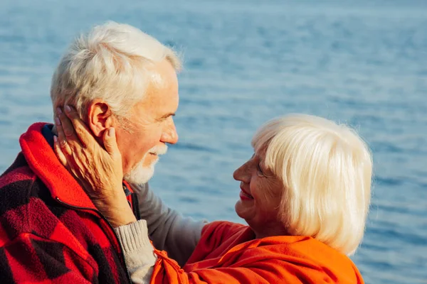 Cheerful senior citizens woman and man are standing and hugging on the lake, against the background of the bridge. — Stock Fotó