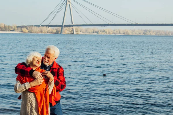 cheerful senior citizens woman and man are standing and hugging on the lake, against the background of the bridge.