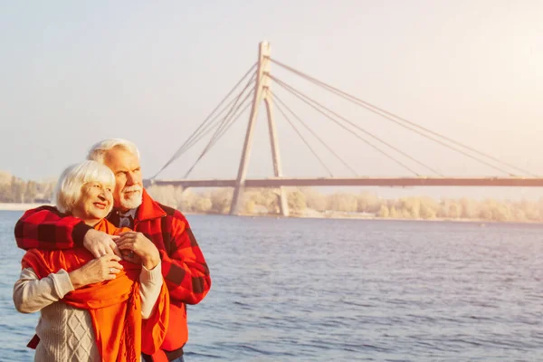 Cheerful senior citizens woman and man are standing and hugging on the lake, against the background of the bridge. — Stock Photo, Image