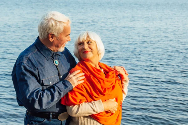 Cheerful senior citizens woman and man are standing and hugging on the lake, against the background of the bridge. — Stock Photo, Image