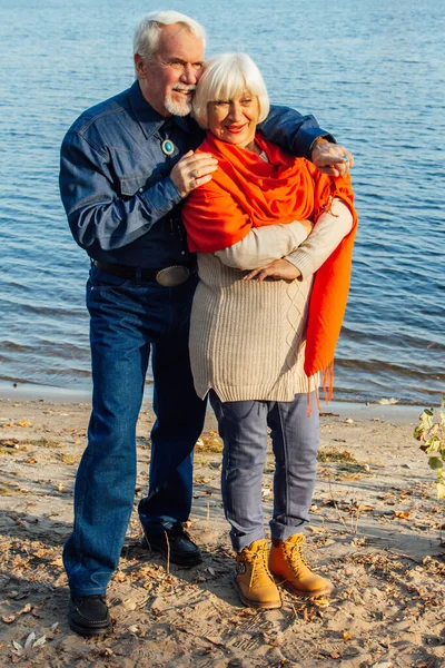 Весела старші громадяни жінка і чоловік стоять і обіймаються на озері, на тлі мосту . — стокове фото