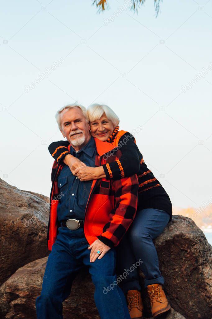 Cheerful elderly people a woman and a man are sitting on the stones and hugging on the lake, against the background of the bridge.