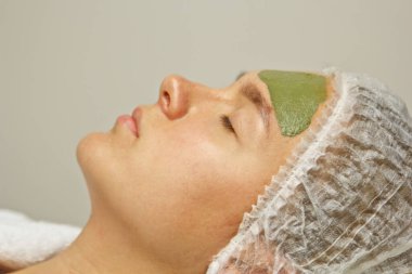 The concept of cosmetology. Close-up of a beautician applying a green mask with a brush on a woman's face. clipart
