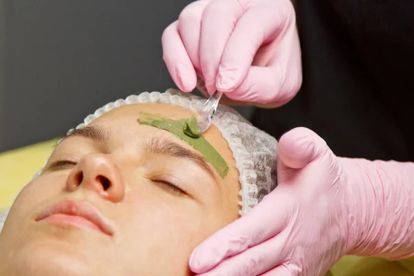 Понятие косметологии. Крупный план косметолога, надевающего зеленую маску с кистью на лицо женщины . — стоковое фото
