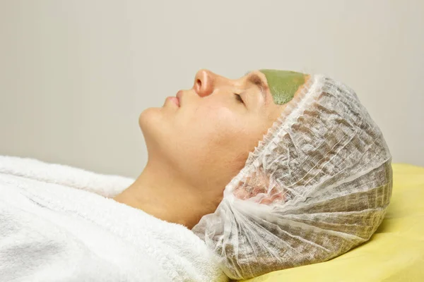 Das Konzept der Kosmetologie. Nahaufnahme einer Kosmetikerin, die eine grüne Maske mit einem Pinsel auf das Gesicht einer Frau aufträgt. — Stockfoto