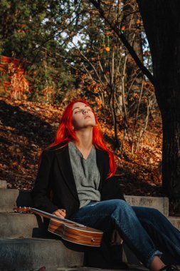 Muhteşem uzun saçlı, seksi, kızıl saçlı kız. Ukulele çalıyor, parkta merdivenlerde oturuyor. Mükemmel bir kadın portresi. Müzik eğitimi, sokak sanatçısı..