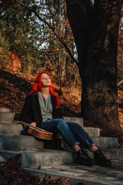 Muhteşem uzun saçlı, seksi, kızıl saçlı kız. Ukulele çalıyor, parkta merdivenlerde oturuyor. Mükemmel bir kadın portresi. Müzik eğitimi, sokak sanatçısı..