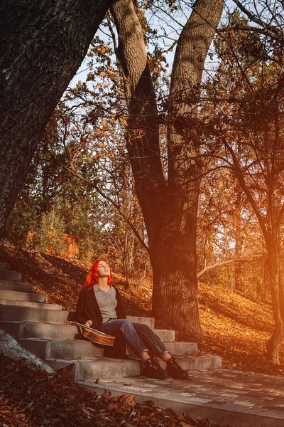 Sexy mooi roodharig meisje met prachtig lang haar. Ukulele aan het spelen, zittend op de trappen in het park. Perfect vrouwenportret. Muziekonderwijs, straatartiest. — Stockfoto