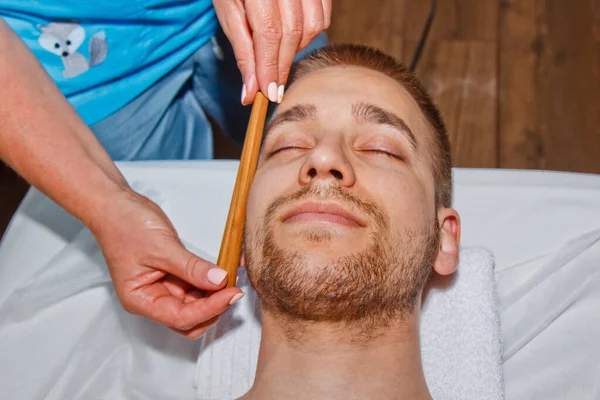 Conceito de bem-estar. Massagista profissional dá a um homem uma massagem facial tailandesa com paus especiais em um centro de spa. Relaxamento e recuperação após o trabalho . — Fotografia de Stock