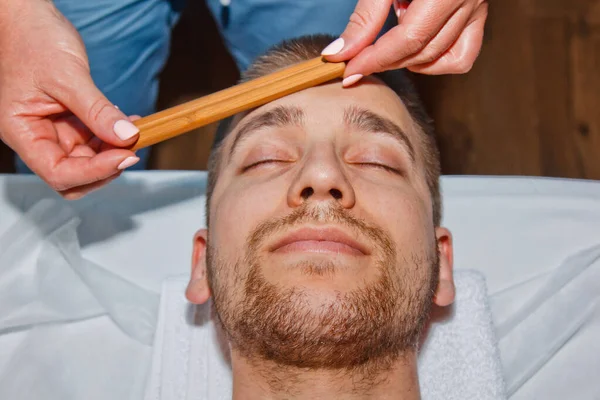 Conceito de bem-estar. Massagista profissional dá a um homem uma massagem facial tailandesa com paus especiais em um centro de spa. Relaxamento e recuperação após o trabalho . — Fotografia de Stock