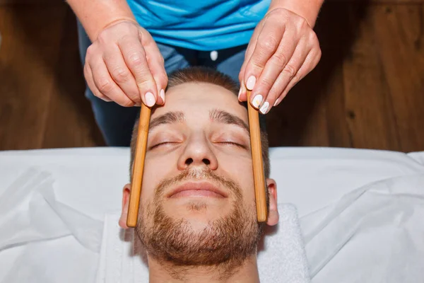 Conceito de bem-estar. Massagista profissional dá a um homem uma massagem facial tailandesa com paus especiais em um centro de spa. Relaxamento e recuperação após o trabalho . — Fotografia de Stock