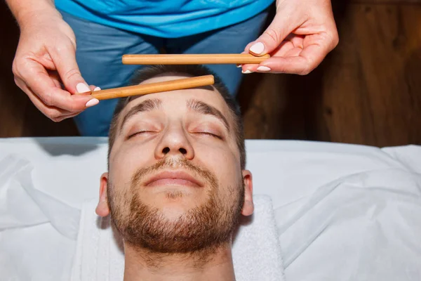 Conceito de bem-estar. Massagista profissional dá a um homem uma massagem facial tailandesa com paus especiais em um centro de spa. Relaxamento e recuperação após o trabalho . — Fotografia de Stock