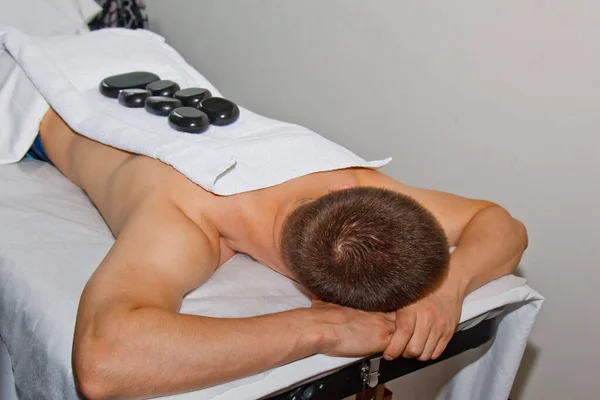 Wellnessconcept. Knappe jongeman ontspannen onder het stimulerende effect van een traditionele hot stone massage in een luxe spa en wellness centrum. Herstel na het werk. — Stockfoto