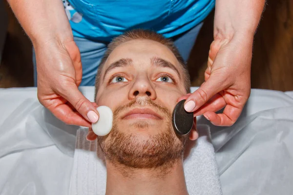 Conceito de bem-estar. Bonito jovem relaxante sob o efeito estimulante de uma massagem facial de pedra quente tradicional em um luxuoso spa e centro de bem-estar. Recuperação após o trabalho . — Fotografia de Stock