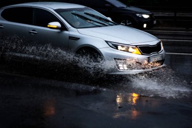 Ukrayna. Kiev - 05,12,2019 Islak şehir asfalt yolunda ilerleyen bir aracın tekerleklerinden su püskürtüyor. Islak bir arabanın tekerleği sular altındaki bir şehir yolunda yağmur altında su birikintisi boyunca hızla hareket eder..