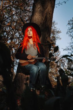 Kızıl saçlı, uzun saçlı kız parkta ukulele çalıyor. Okul, müzik eğitimi konsepti, öğrenci telli çalgılar çalmayı öğreniyor. Bir müzisyenin elleri, klasik, melodi, yaratıcılık.