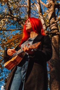 Kızıl saçlı, uzun saçlı kız parkta ukulele çalıyor. Okul, müzik eğitimi konsepti, öğrenci telli çalgılar çalmayı öğreniyor. Bir müzisyenin elleri, klasik, melodi, yaratıcılık.