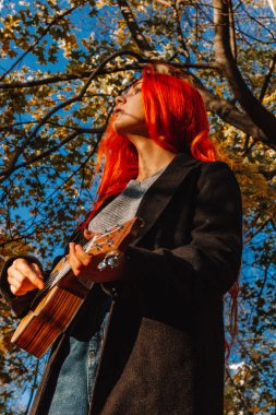 Kızıl saçlı, uzun saçlı kız parkta ukulele çalıyor. Okul, müzik eğitimi konsepti, öğrenci telli çalgılar çalmayı öğreniyor. Bir müzisyenin elleri, klasik, melodi, yaratıcılık.