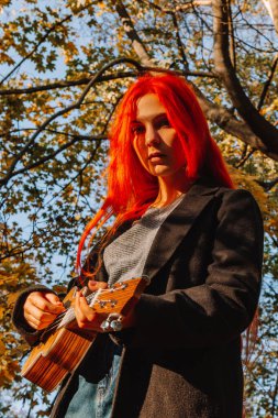 Kızıl saçlı, uzun saçlı kız parkta ukulele çalıyor. Okul, müzik eğitimi konsepti, öğrenci telli çalgılar çalmayı öğreniyor. Bir müzisyenin elleri, klasik, melodi, yaratıcılık.