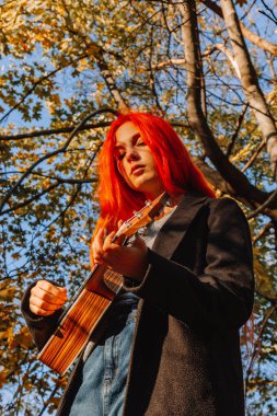 Kızıl saçlı, uzun saçlı kız parkta ukulele çalıyor. Okul, müzik eğitimi konsepti, öğrenci telli çalgılar çalmayı öğreniyor. Bir müzisyenin elleri, klasik, melodi, yaratıcılık.