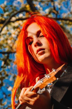 Kızıl saçlı, uzun saçlı kız parkta ukulele çalıyor. Okul, müzik eğitimi konsepti, öğrenci telli çalgılar çalmayı öğreniyor. Bir müzisyenin elleri, klasik, melodi, yaratıcılık.