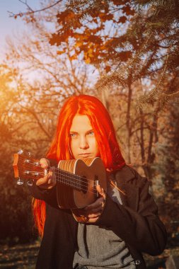 Kızıl saçlı, uzun saçlı kız parkta ukulele çalıyor. Okul, müzik eğitimi konsepti, öğrenci telli çalgılar çalmayı öğreniyor. Bir müzisyenin elleri, klasik, melodi, yaratıcılık.