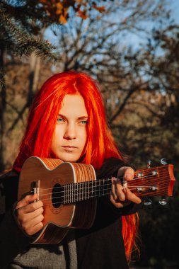 Kızıl saçlı, uzun saçlı kız parkta ukulele çalıyor. Okul, müzik eğitimi konsepti, öğrenci telli çalgılar çalmayı öğreniyor. Bir müzisyenin elleri, klasik, melodi, yaratıcılık.