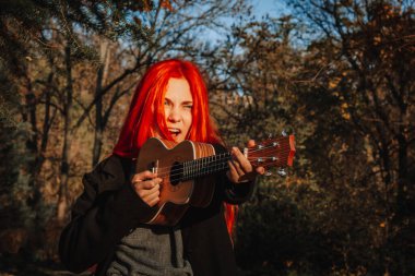 Kızıl saçlı, uzun saçlı kız parkta ukulele çalıyor. Okul, müzik eğitimi konsepti, öğrenci telli çalgılar çalmayı öğreniyor. Bir müzisyenin elleri, klasik, melodi, yaratıcılık.