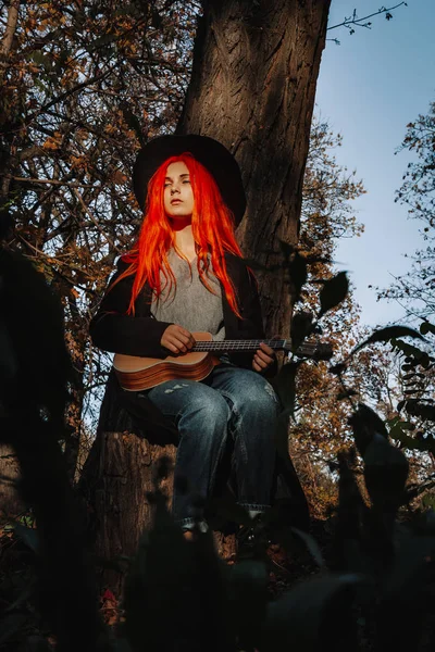 Kızıl saçlı, uzun saçlı kız parkta ukulele çalıyor. Okul, müzik eğitimi konsepti, öğrenci telli çalgılar çalmayı öğreniyor. Bir müzisyenin elleri, klasik, melodi, yaratıcılık.
