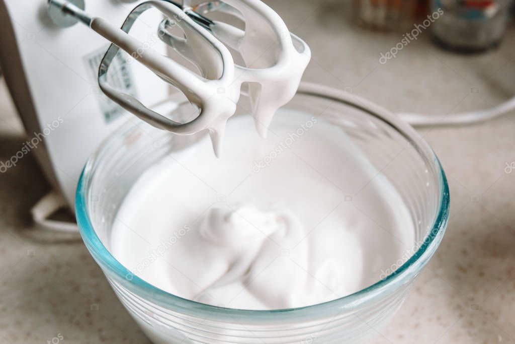 Baking concept. Whipped cream of egg whites for perfect peaks in a glass bowl, with a mixer on a marble white table. Meringue cookie recipe.