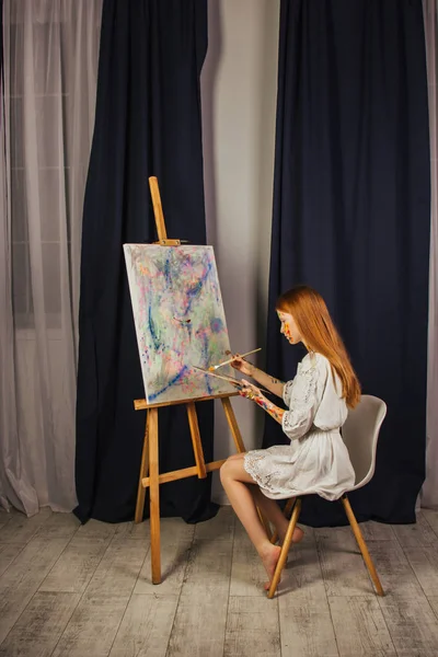 La joven de la artista en un vestido blanco claro, pinta un cuadro sobre lienzo en el taller. La cara está manchada con pinturas. Un joven estudiante usa pinceles, lienzos y caballetes. Trabajos creativos . —  Fotos de Stock