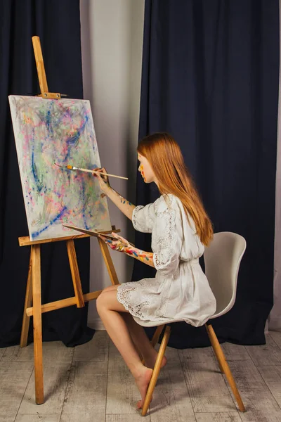 The young girl of the artist in a light white dress, paints a picture on canvas in the workshop. The face is stained with paints. A young student uses brushes, canvases and easels. Creative work.