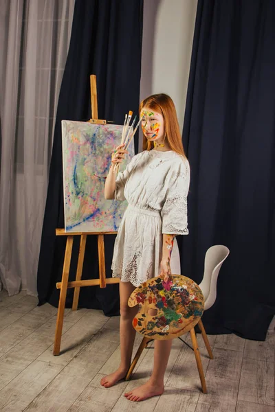 A művész fiatal lánya világos fehér ruhában vászonra fest egy képet a műhelyben. Az arcán festékek vannak. Egy fiatal diák ecseteket, vásznakat és festőállványokat használ. Alkotói munka. — Stock Fotó