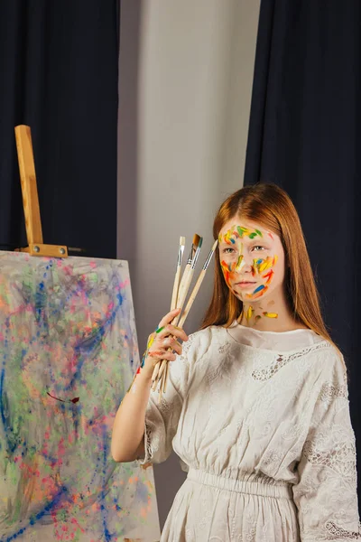 La joven de la artista en un vestido blanco claro, pinta un cuadro sobre lienzo en el taller. La cara está manchada con pinturas. Un joven estudiante usa pinceles, lienzos y caballetes. Trabajos creativos . — Foto de Stock