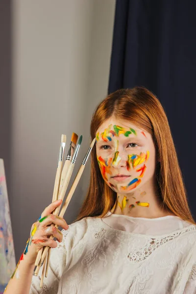 The young girl of the artist in a light white dress, paints a picture on canvas in the workshop. The face is stained with paints. A young student uses brushes, canvases and easels. Creative work. — Stock Photo, Image