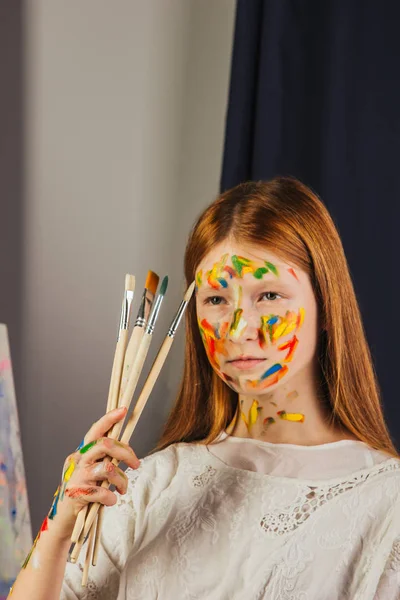 Mladá dívka umělce v světle bílých šatech, maluje obraz na plátno v dílně. Obličej je potřísněn barvami. Mladý student používá štětce, plátna a lehátka. Tvůrčí práce. — Stock fotografie