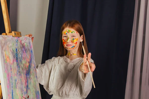 La joven de la artista en un vestido blanco claro, pinta un cuadro sobre lienzo en el taller. La cara está manchada con pinturas. Un joven estudiante usa pinceles, lienzos y caballetes. Trabajos creativos . — Foto de Stock