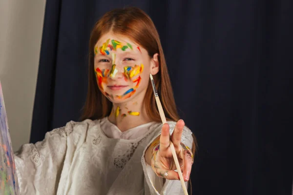 Η νεαρή κοπέλα του καλλιτέχνη με ένα λευκό ανοιχτό φόρεμα, ζωγραφίζει μια εικόνα σε καμβά στο εργαστήριο. Το πρόσωπο είναι βαμμένο με χρώματα. Ένας νεαρός μαθητής χρησιμοποιεί βούρτσες, καμβάδες και καβαλίνες. Δημιουργική εργασία. — Φωτογραφία Αρχείου