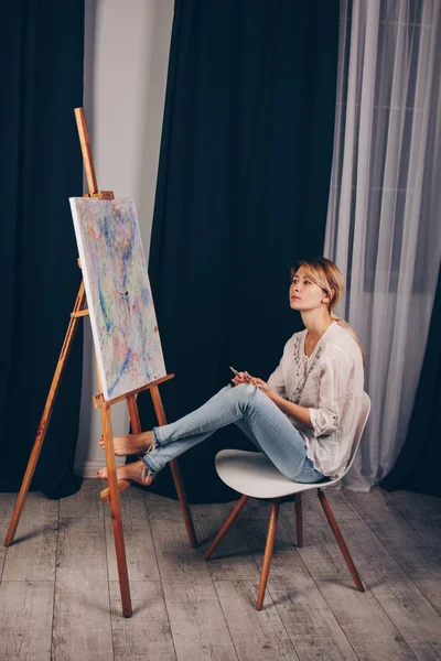 Young artist girl in a white shirt and blue jeans, paints a picture on canvas in the workshop. A young student uses brushes, canvases and easels. Creative work for children and adults. — ストック写真