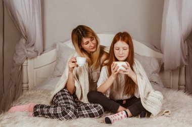 Yeni yıl 2020. Anne ve kızıl saçlı kız yatakta oturmuş köpüklü kakao içiyorlar. Battaniyeyle örtülü, sarıl.