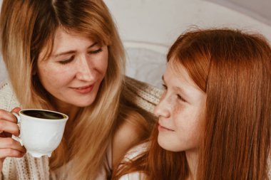 Yeni yıl 2020. Anne ve kızıl saçlı kız yatakta oturmuş köpüklü kakao içiyorlar. Battaniyeyle örtülü, sarıl.