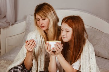 Yeni yıl 2020. Anne ve kızıl saçlı kız yatakta oturmuş köpüklü kakao içiyorlar. Battaniyeyle örtülü, sarıl.