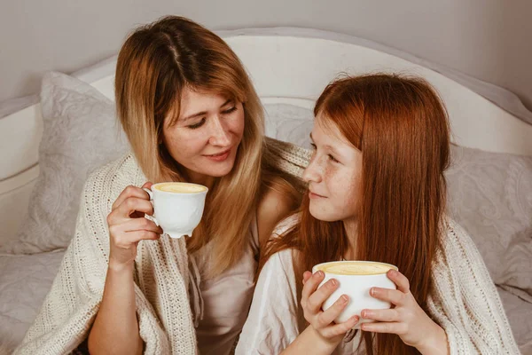 Новый 2020 год. Мама и рыжая дочь сидят на кровати и пьют какао с пеной. Покрытые одеялом, обнимашки . — стоковое фото