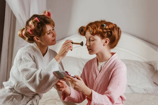 Happy family. Young beautiful mother does makeup of her beautiful daughter on the bed in the bedroom. The red-haired daughter with curlers on her head is laughing.